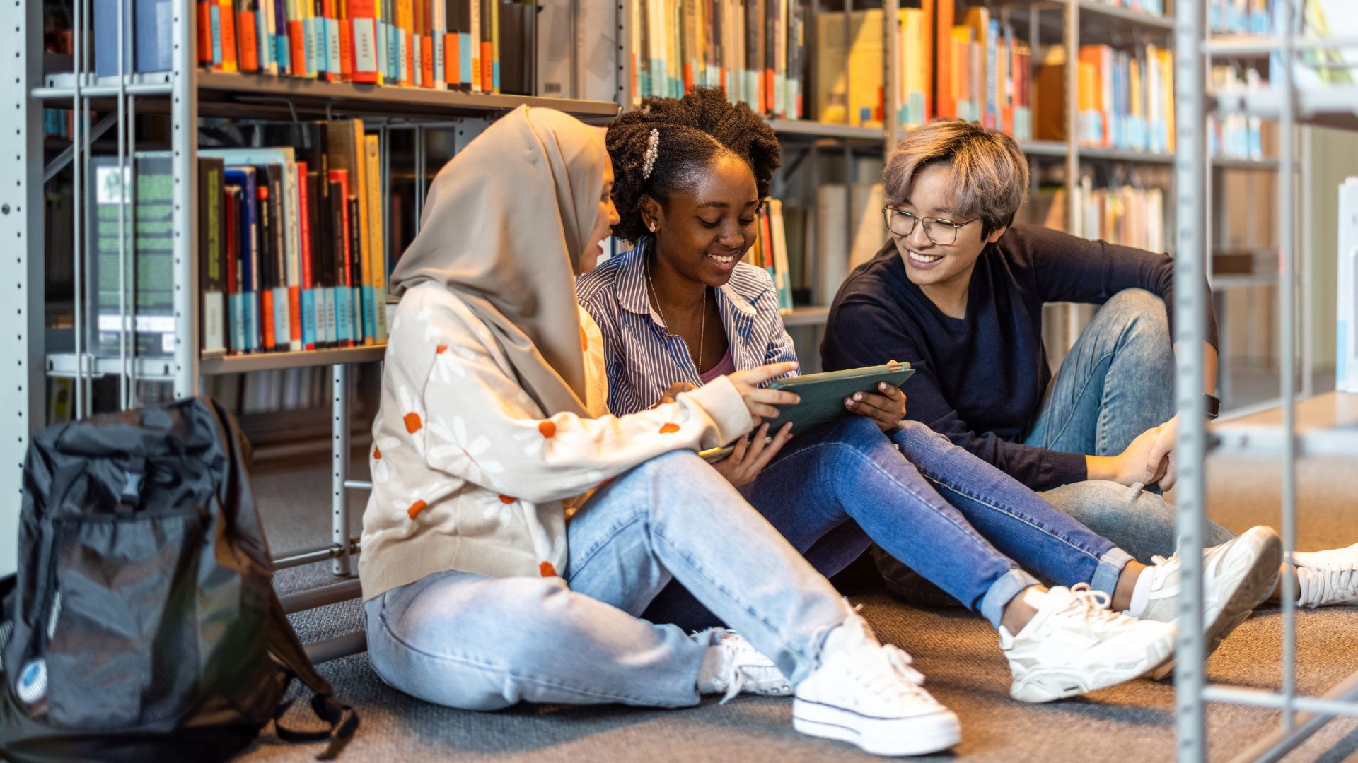 Three individuals sit at the base of a bookshelf, looking at a tablet