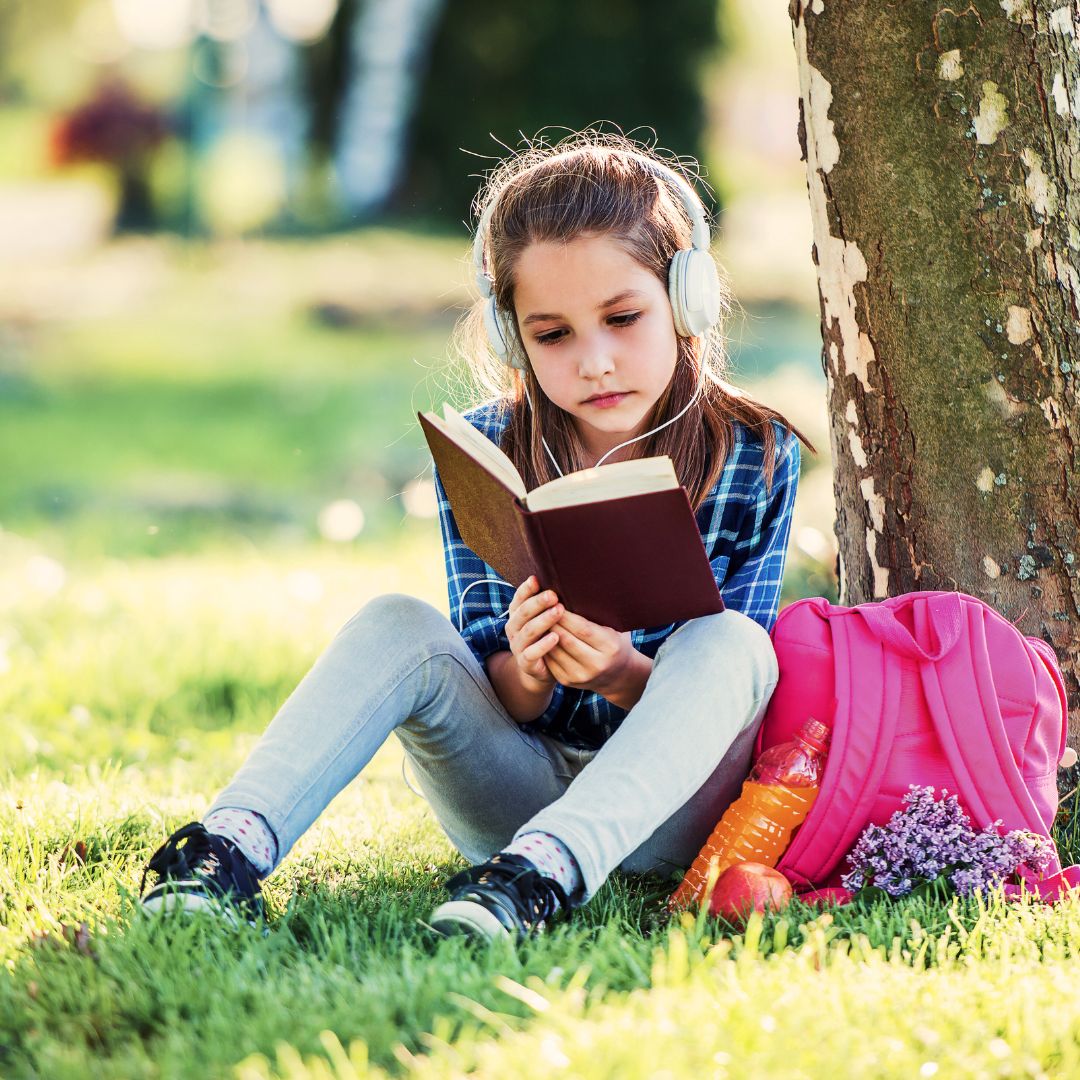 A child under a tree reads a physical book with headphones on.