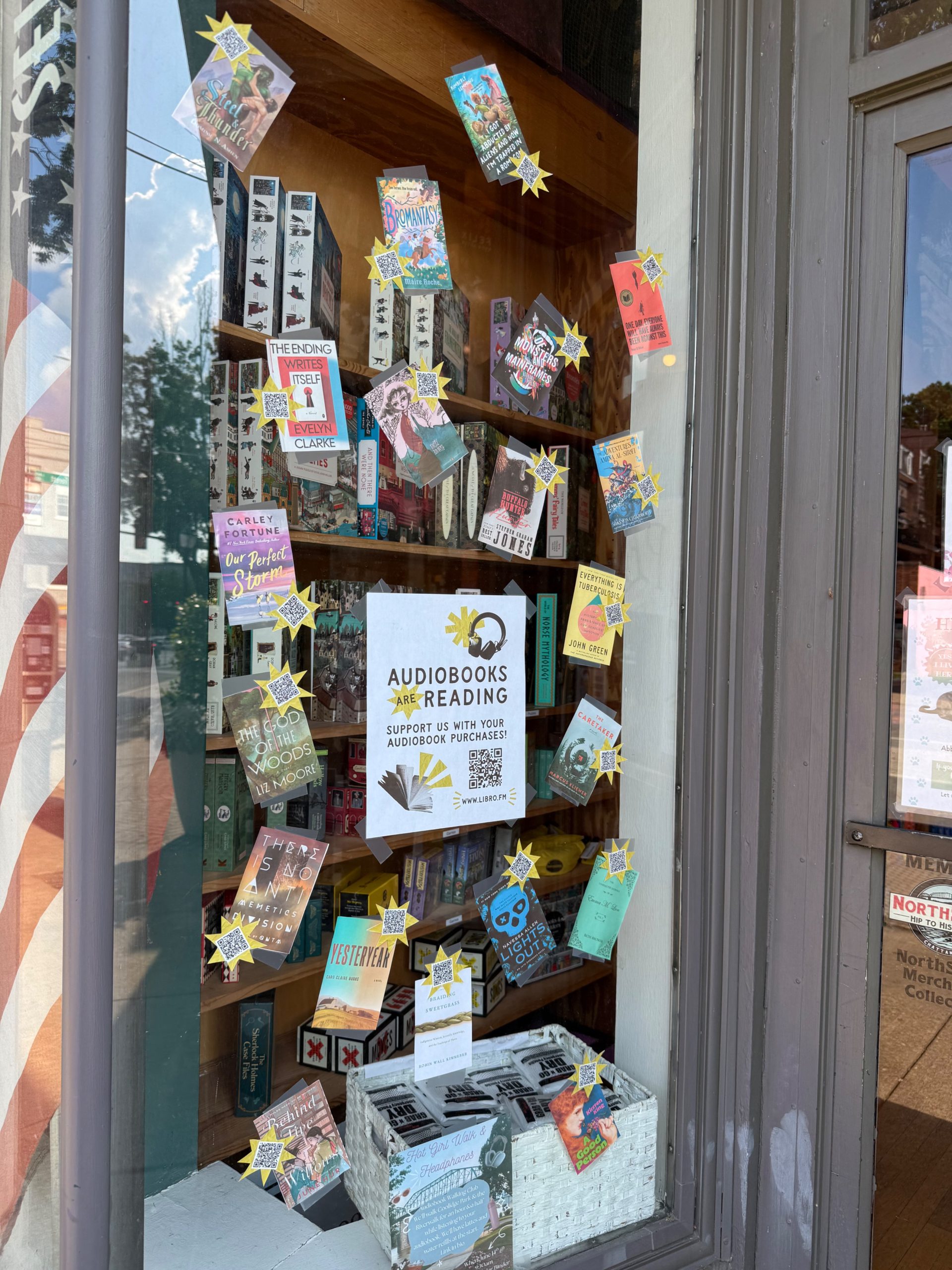 Storefront window decorated with audiobook cover images and QR codes, and a sign that reads "Audiobooks are reading. Support us with your audiobook purchases."