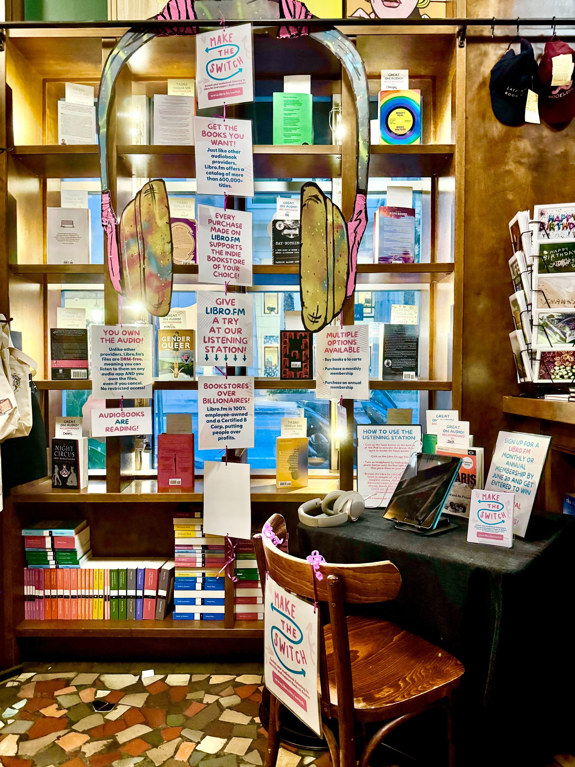 Audiobook listening station with hand-painted giant headphone cutout, signs highlighting benefits of listening to audiobooks on Libro.fm, and a table with headphones and a tablet.