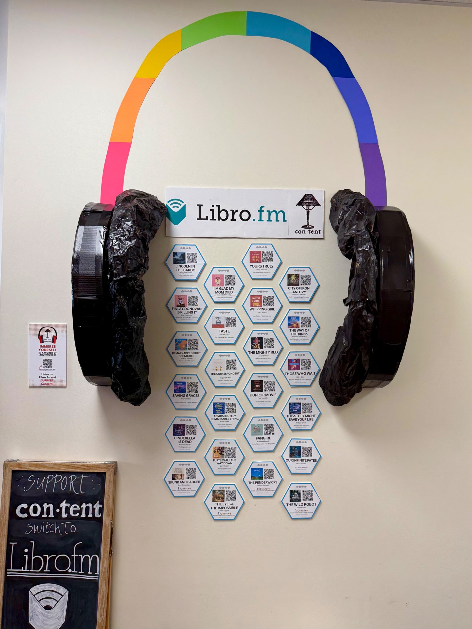 Audiobook display featuring a giant headphone installation and hexagon-shaped paper cards showing audiobook covers and QR codes