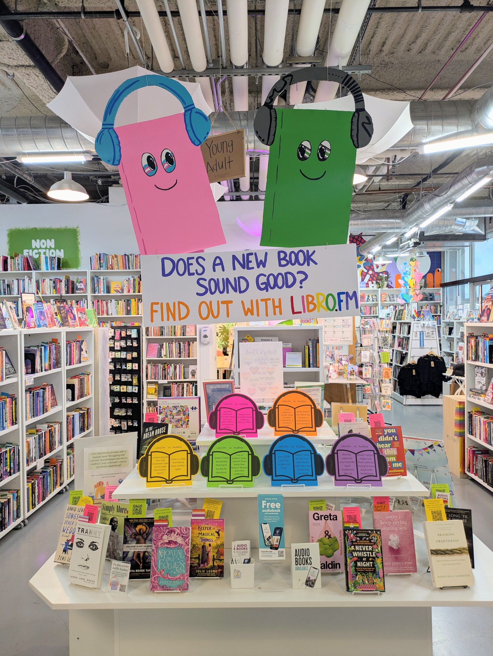 Audiobook display featuring two hand-drawn audiobook mascots and cardboard sign reading "Does a new book sound good? Find out with Libro.fm", and a table of print books with bookmarks that include audiobook QR codes.