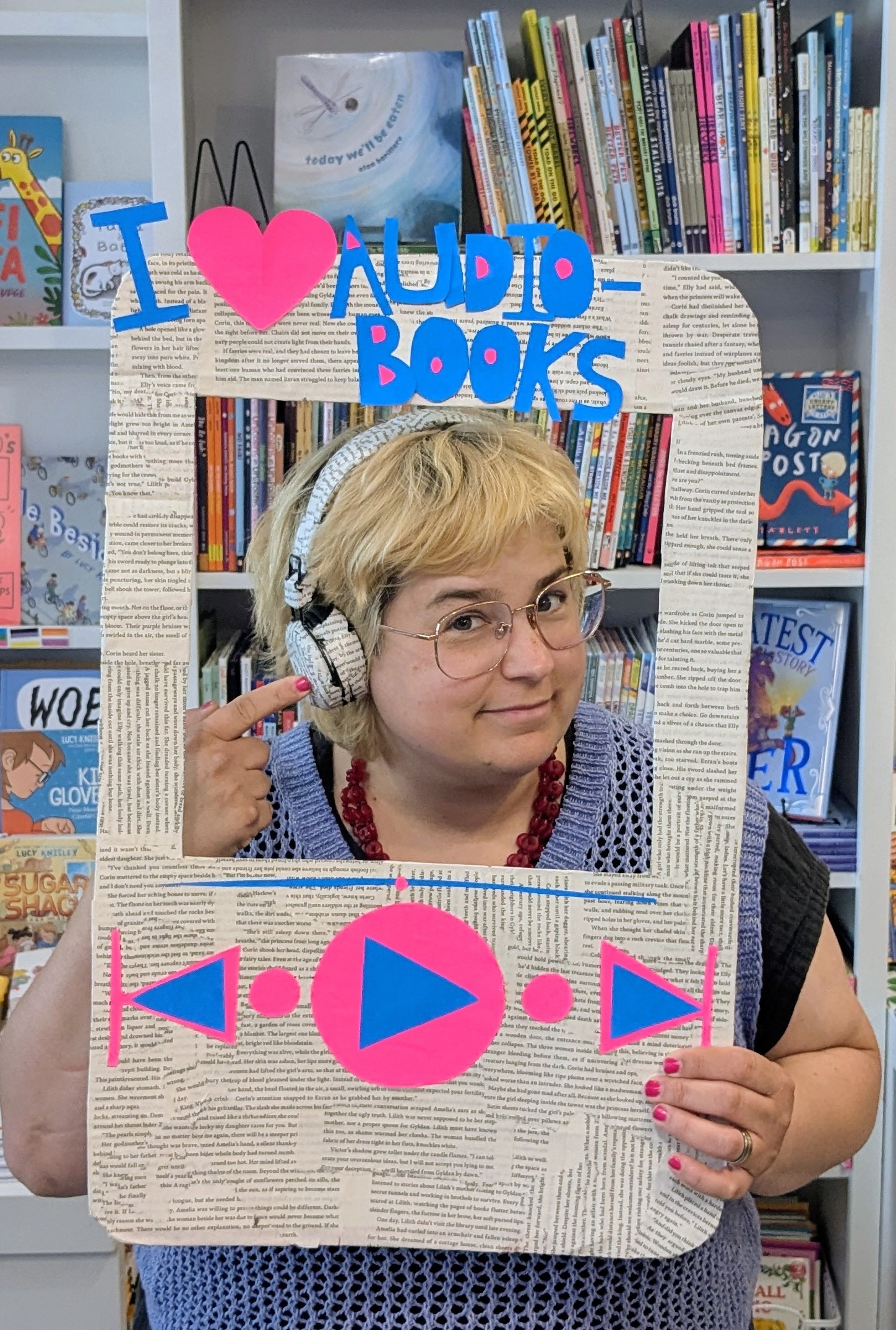 Person posing with book page-decorated headphones and holding a handcraft "I Love Audiobooks" sign covered made from book-page paper.