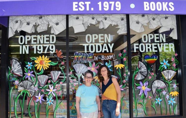 Two people, booksellers at Women & Children First in Chicago, IL stand in front of their bookstore, with a window that reads "Opened in 1979; Open today; Open forever."