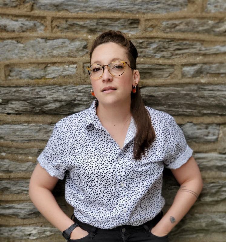 Sara Nović stands in front of a stone wall, with her hands tucked into her pockets. She wears a button down blue and white shirt with tortoiseshell glasses. 
